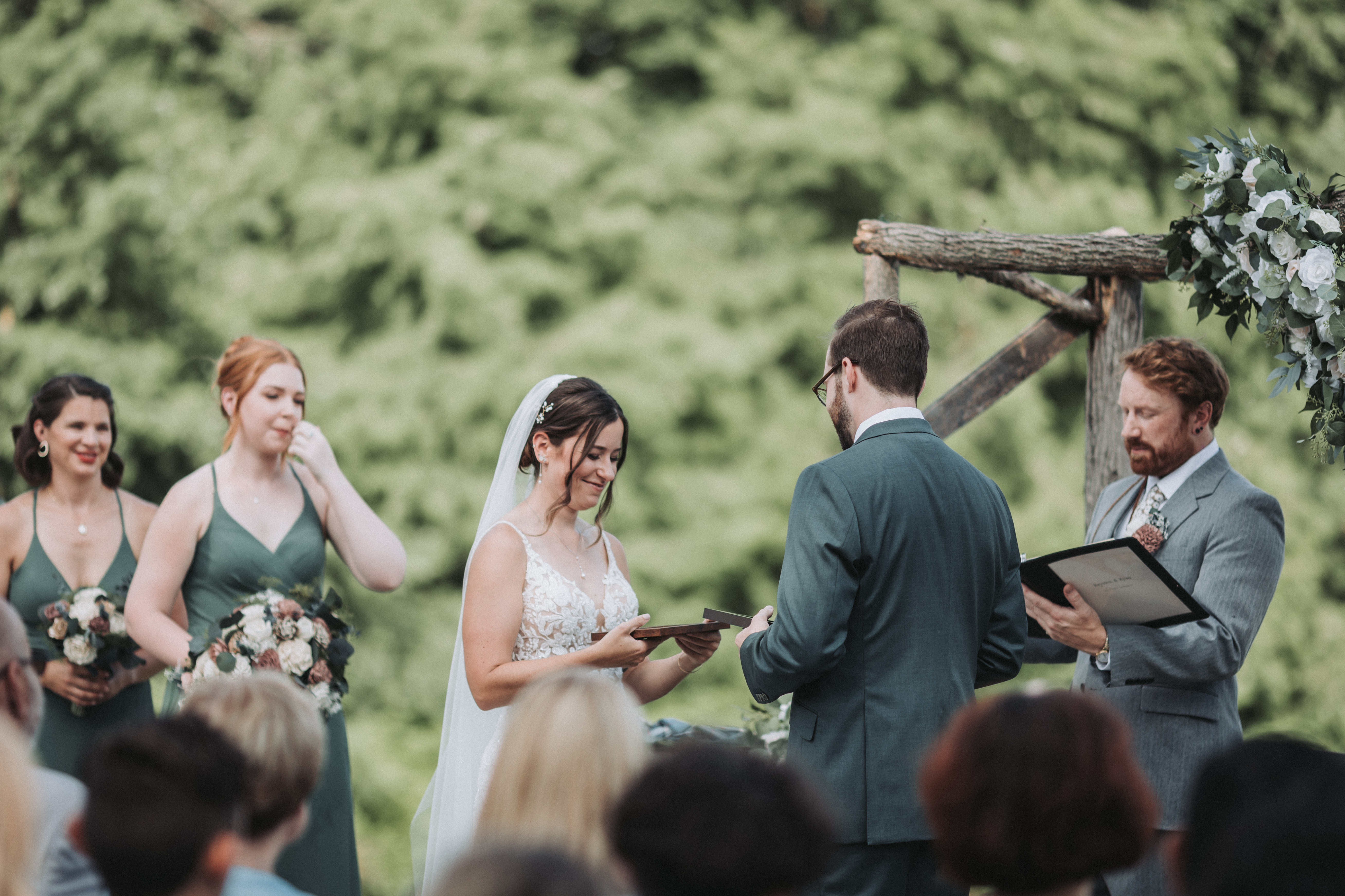 At an outdoor ceremony beneath a rustic log arbor, the bride smiles down at a small wooden keepsake she and the groom are holding while the officiant reads from a black folder and a bridesmaid in green dabs her eye, seen past the heads of seated guests
