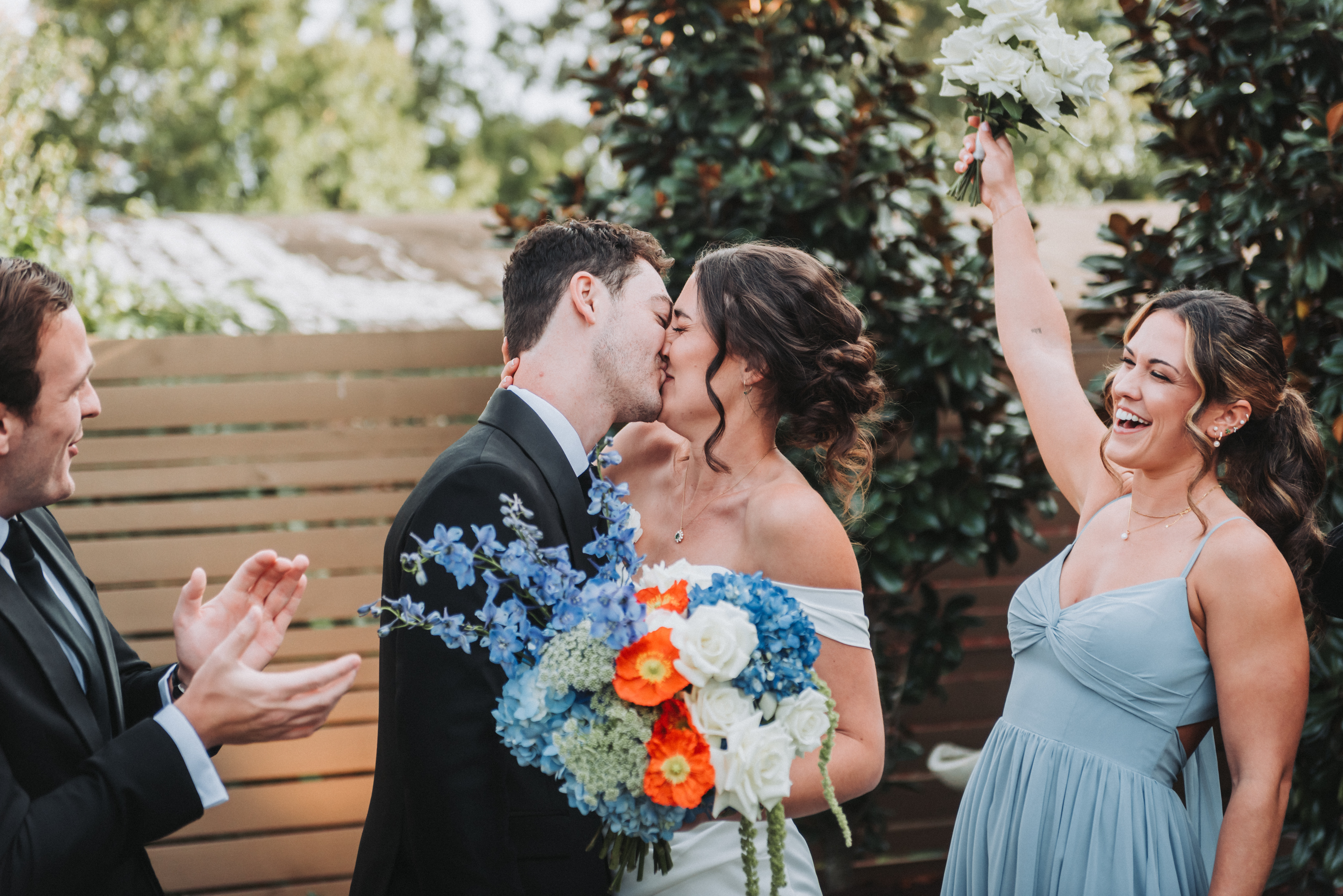 In front of a wooden fence and magnolia leaves, a bride and groom kiss behind her bouquet of blue delphinium and orange poppies, while a groomsman claps and a bridesmaid in pale blue cheers with her bouquet raised