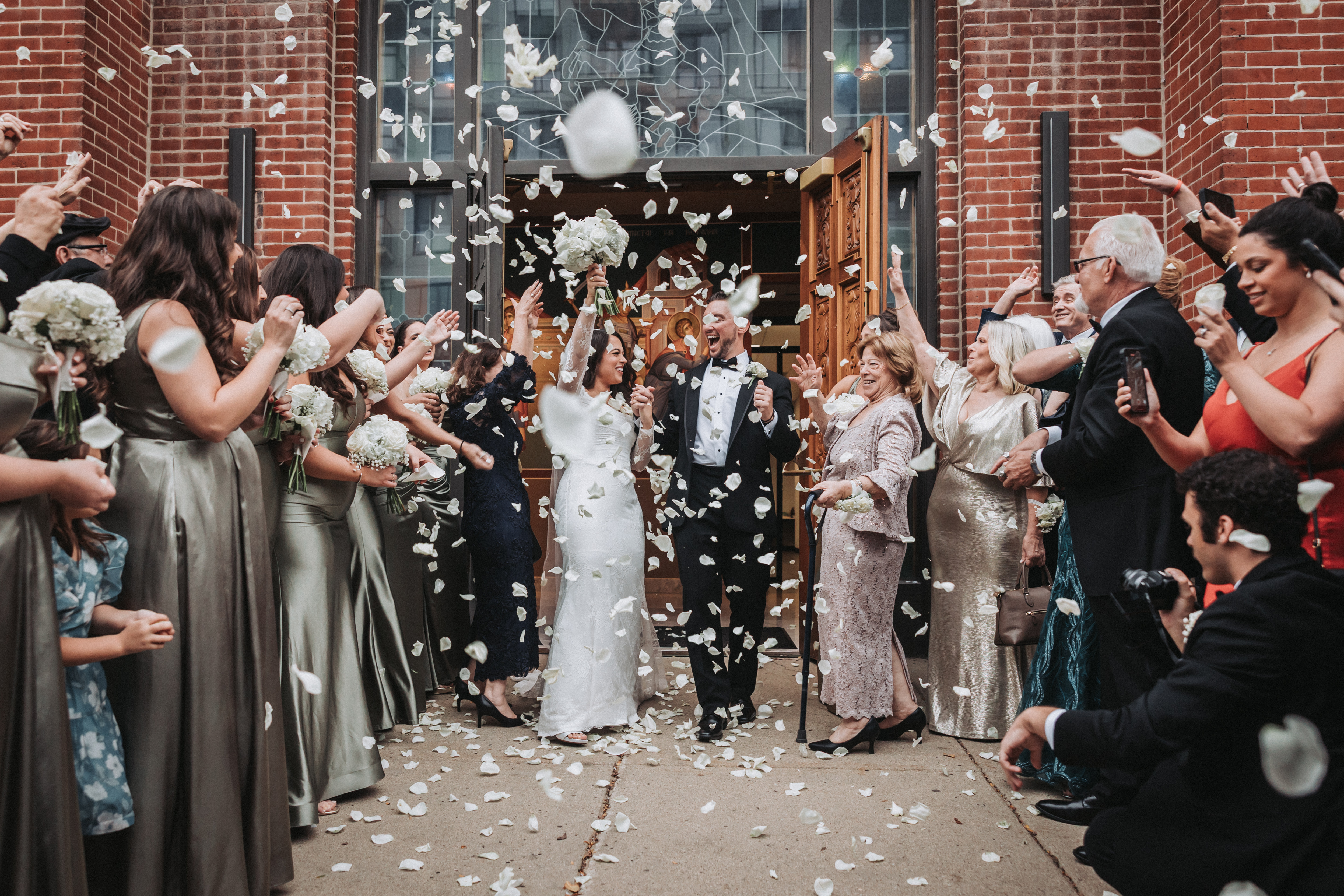 Outside a brick church, guests throw white rose petals as a laughing bride and groom walk out through its carved wooden doors, while guests on the right hold up their phones to film them