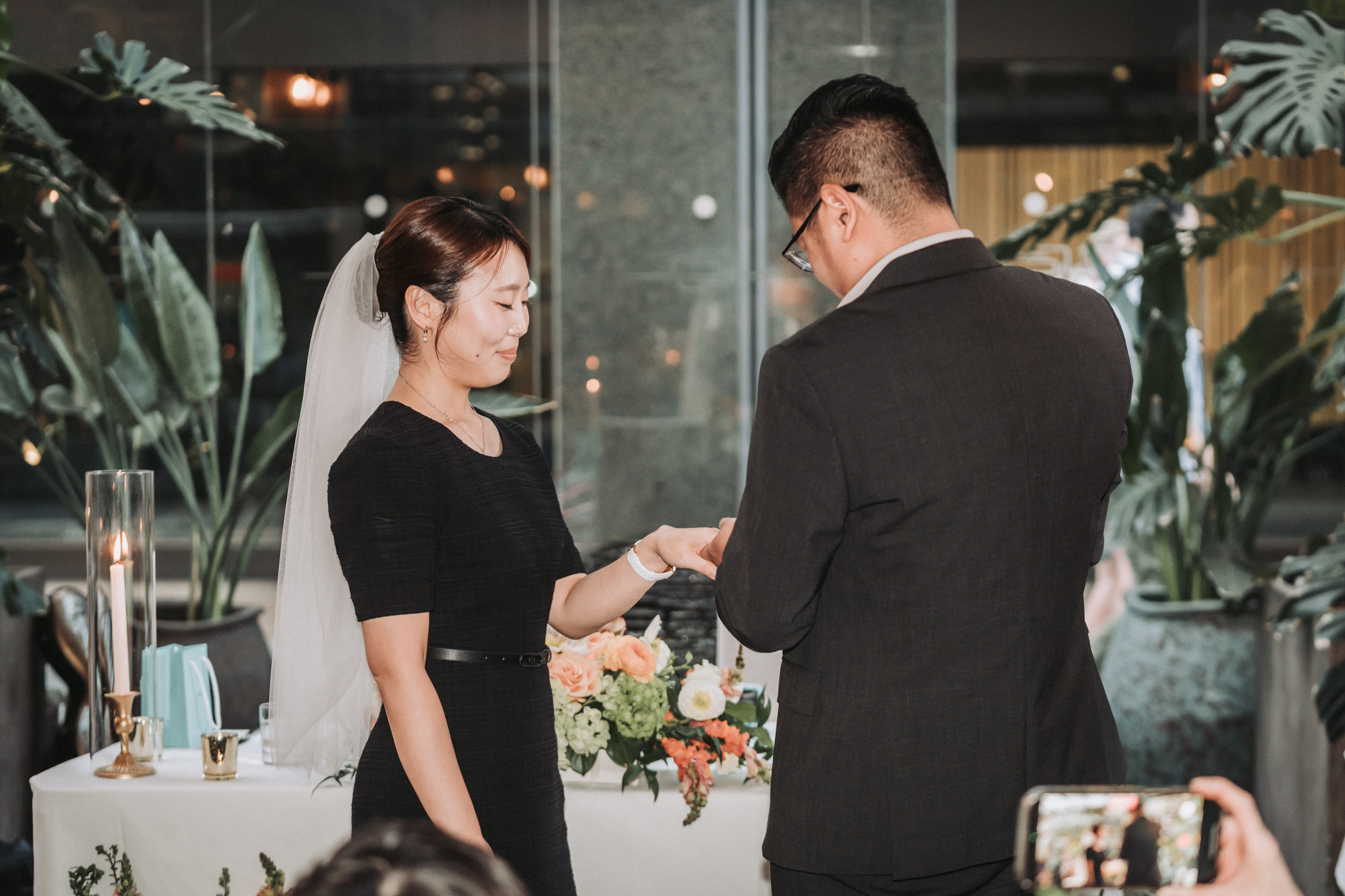 At a small indoor ceremony among potted palms, a bride in a short veil and a black dress smiles as the groom slides a ring onto her finger, while a guest in the foreground films it on a phone