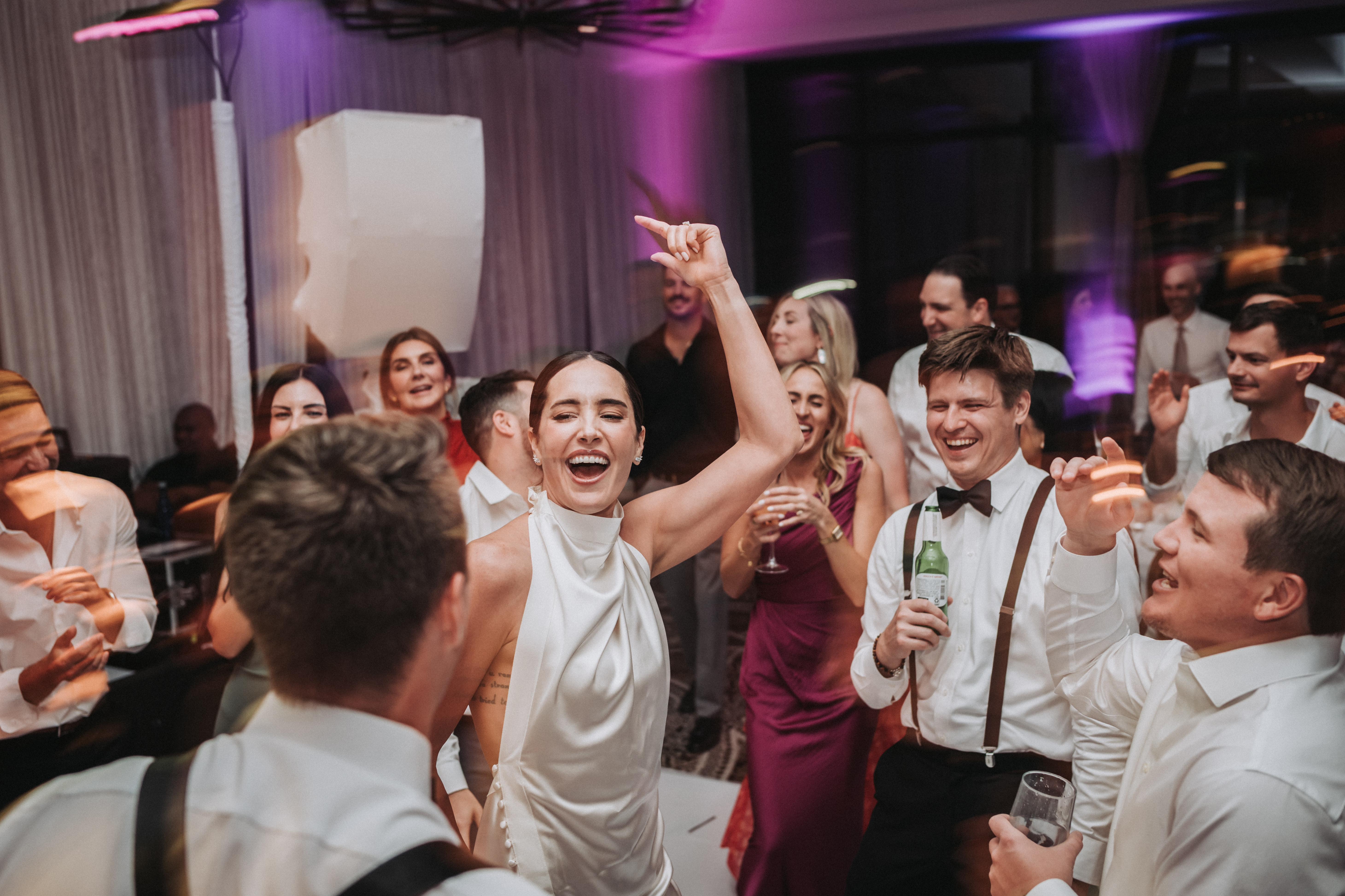 Bride laughing with one arm raised on a crowded dance floor, guests singing around her