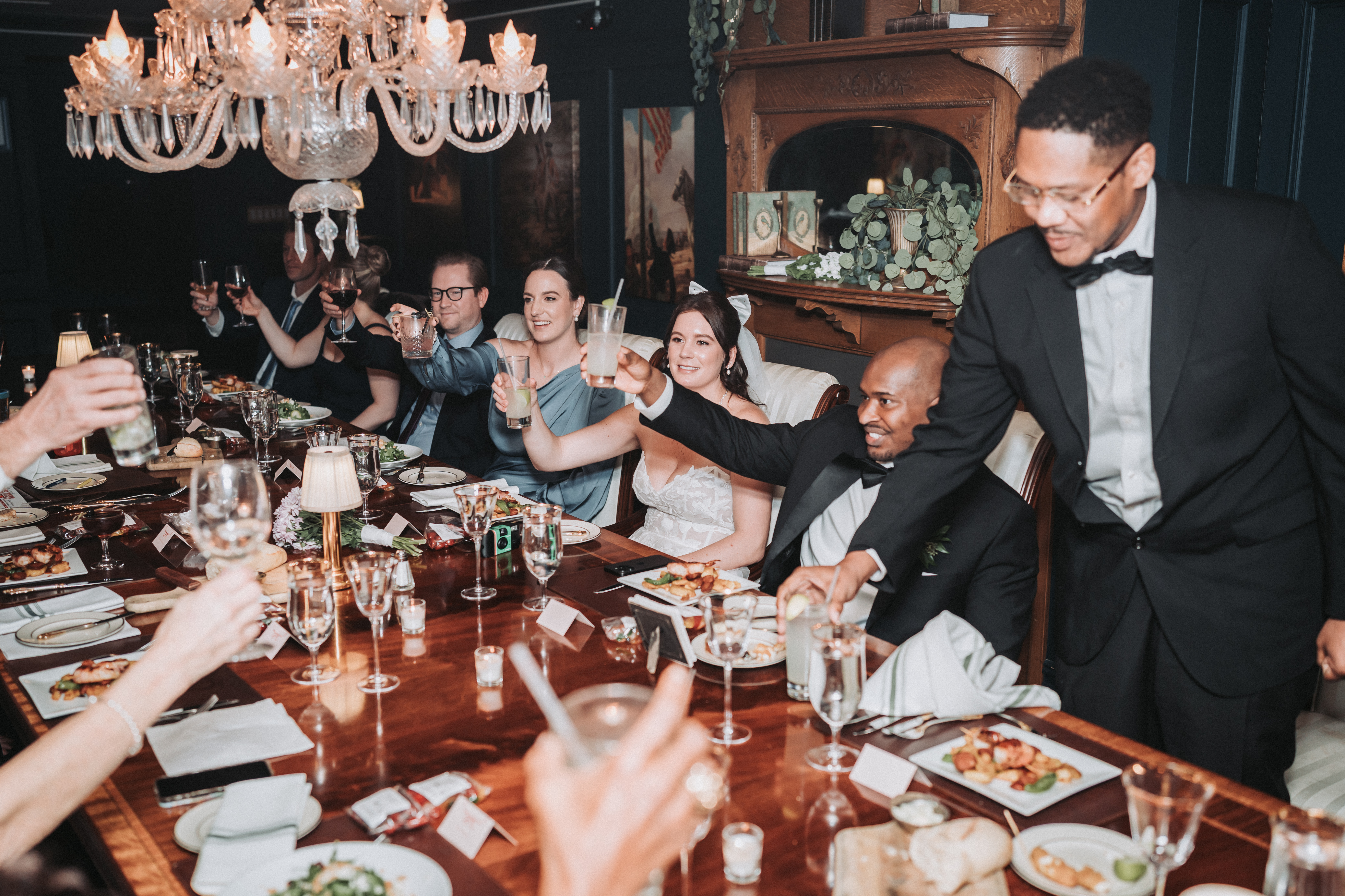 Under a crystal chandelier, a bride and groom and their guests raise their glasses in a toast around a long candlelit dinner table as a guest in a bow tie leans in to clink