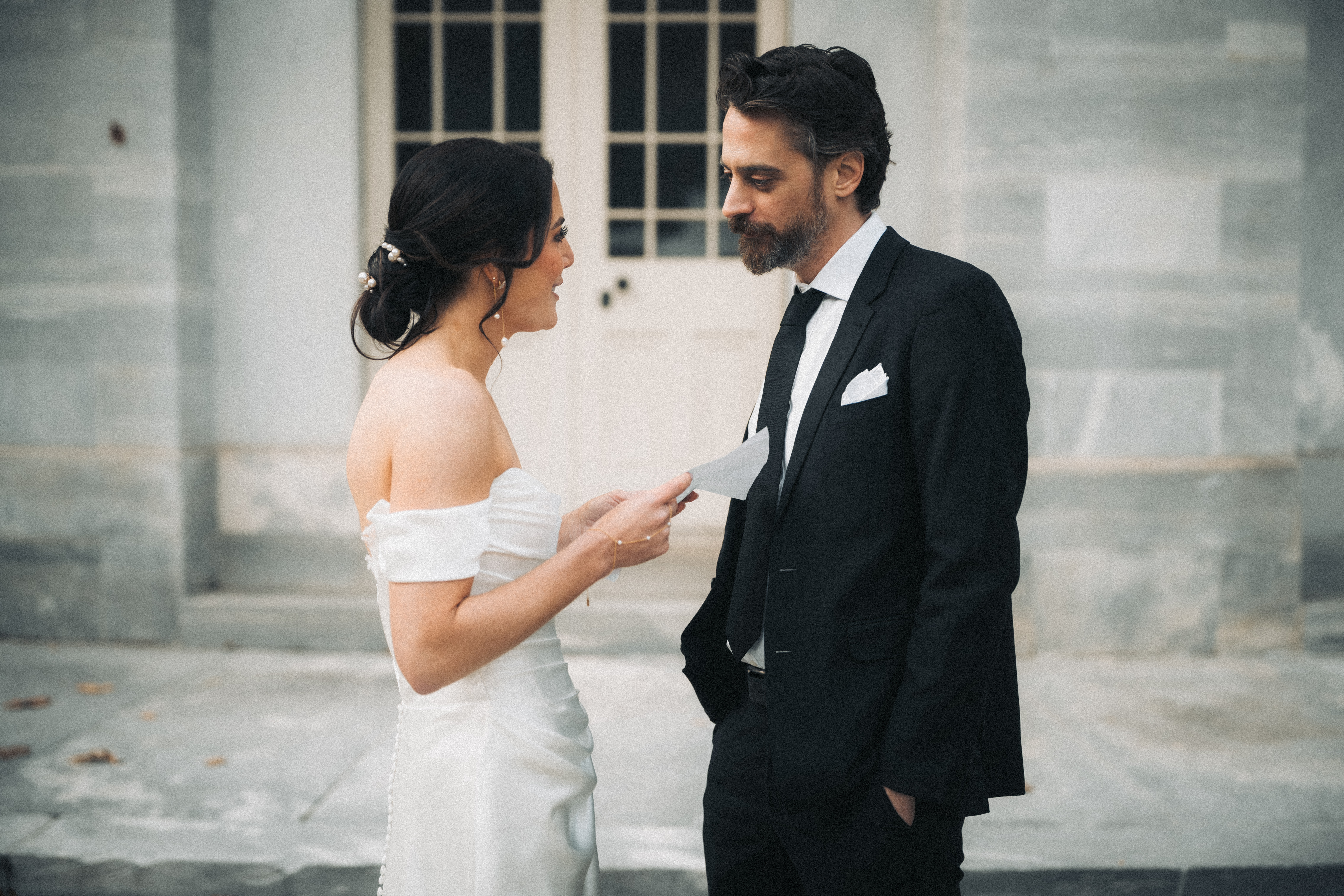 Bride reading her private vows aloud from a folded card while her partner listens, standing outside a stone building