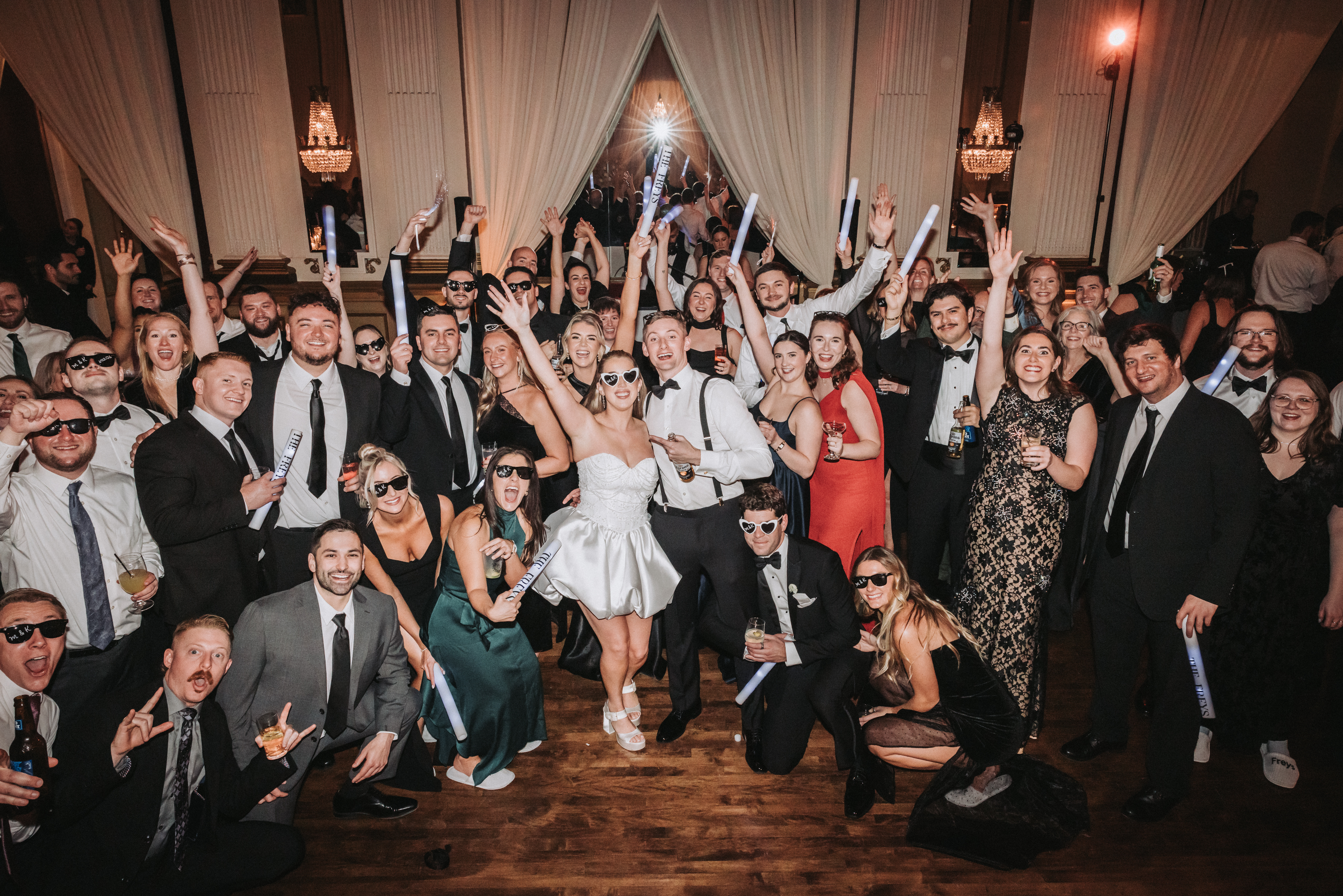 A bride in a short white dress and heart-shaped sunglasses stands in the middle of a crowd of guests on a ballroom dance floor, everyone cheering with arms and glowing foam batons raised