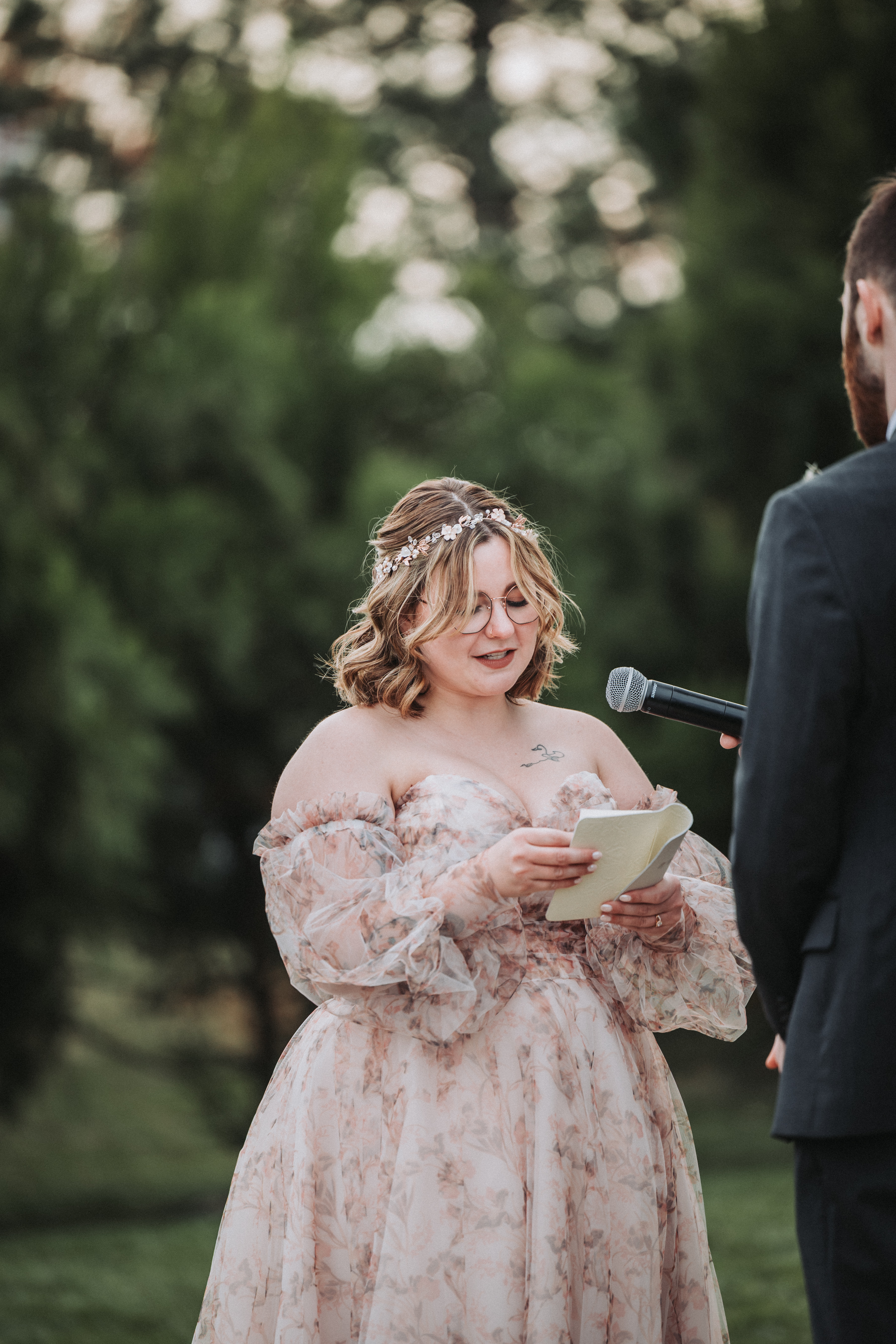 Bride in a floral off-the-shoulder gown reading her vows from a small notebook while the groom holds a microphone out to her